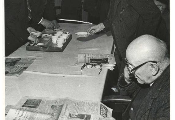 Mannen aan een lange tafel met koffie en kranten.