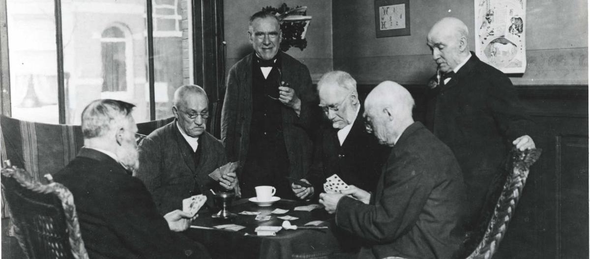 Vier mannen spelen een kaartspel aan tafel, terwijl twee andere mannen toekijken