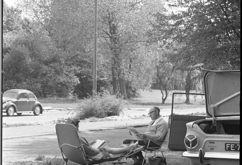 Bermtoerisme: naast een rijweg zitten in bosachtige omgeving een man en vrouw in recreatiestoelen te lezen; naast hun auto met geopende kofferbak en -portier.