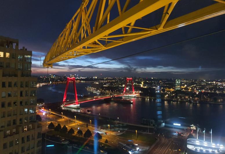 Een opname voor zonsopgang van de verlichte Willemsbrug over de Nieuwe Maas en een blik over de stad in zuidoostelijke richting. Met het horizontale gedeelte van de kraan in beeld