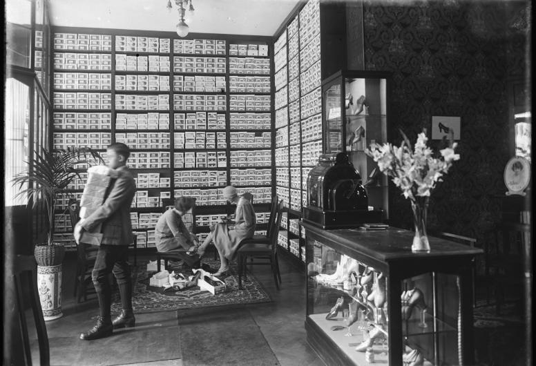 Een foto van het interieur van de schoenenwinkel R.L. Corbeau aan de West-Kruiskade in 1926. Een man verlaat het pand met een stapel pakketjes op zijn arm. Een verkoopmedewerkster helpt een mevrouw bij het aanpassen van een paar hakken.