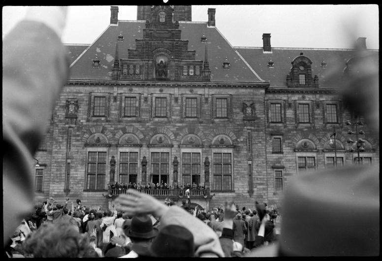 Een foto van het stadhuis genomen vanaf het plein ervoor. Er staat een menigte mensen voor de fotograaf. Mensen zwaaien naar de groep op het bordes. Daar staan burgemeester Oud en Winston Churchill