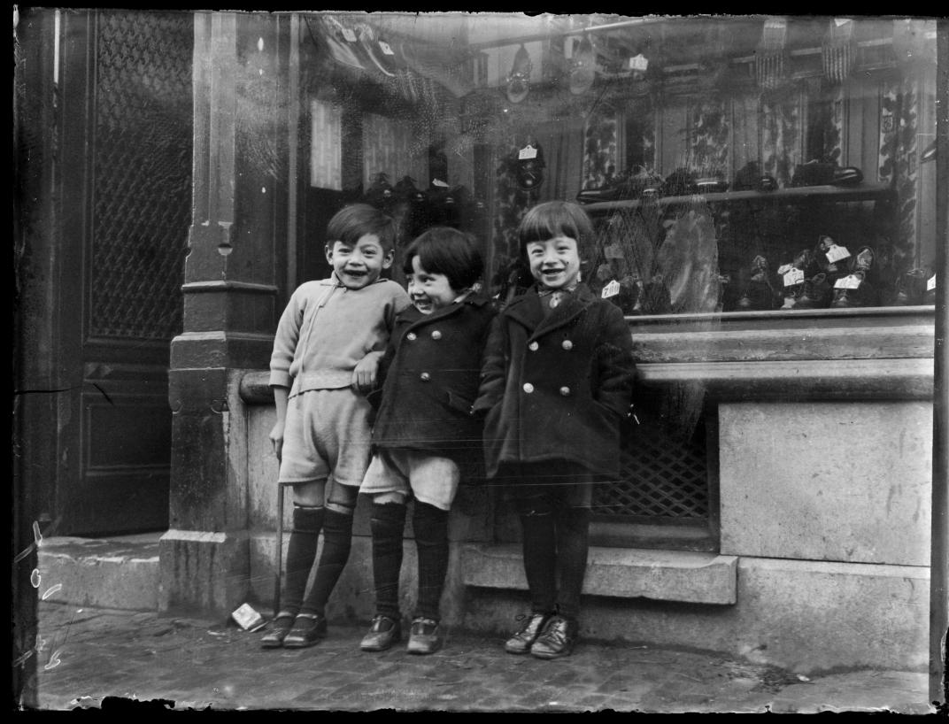 Drie Chinese kinderen staan voor de etalage van een schoenenwinkel, mogelijk in de Delistraat op Katendrecht. Collectie Spaarnestad