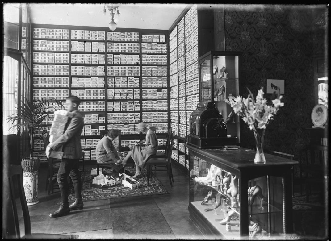 Een foto van het interieur van de schoenenwinkel R.L. Corbeau aan de West-Kruiskade in 1926. Een man verlaat het pand met een stapel pakketjes op zijn arm. Een verkoopmedewerkster helpt een mevrouw bij het aanpassen van een paar hakken.