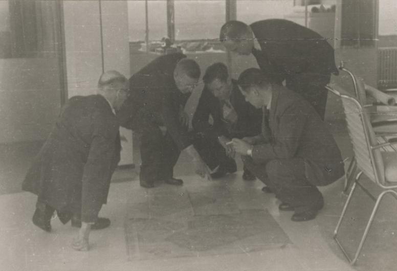 Tijdens een vergadering in de Van Nelle-fabriek in juni 1945 buigen 5 mannen zich over kaarten van Rotterdam die op de grond liggen. Geheel rechts A. van der Goot.